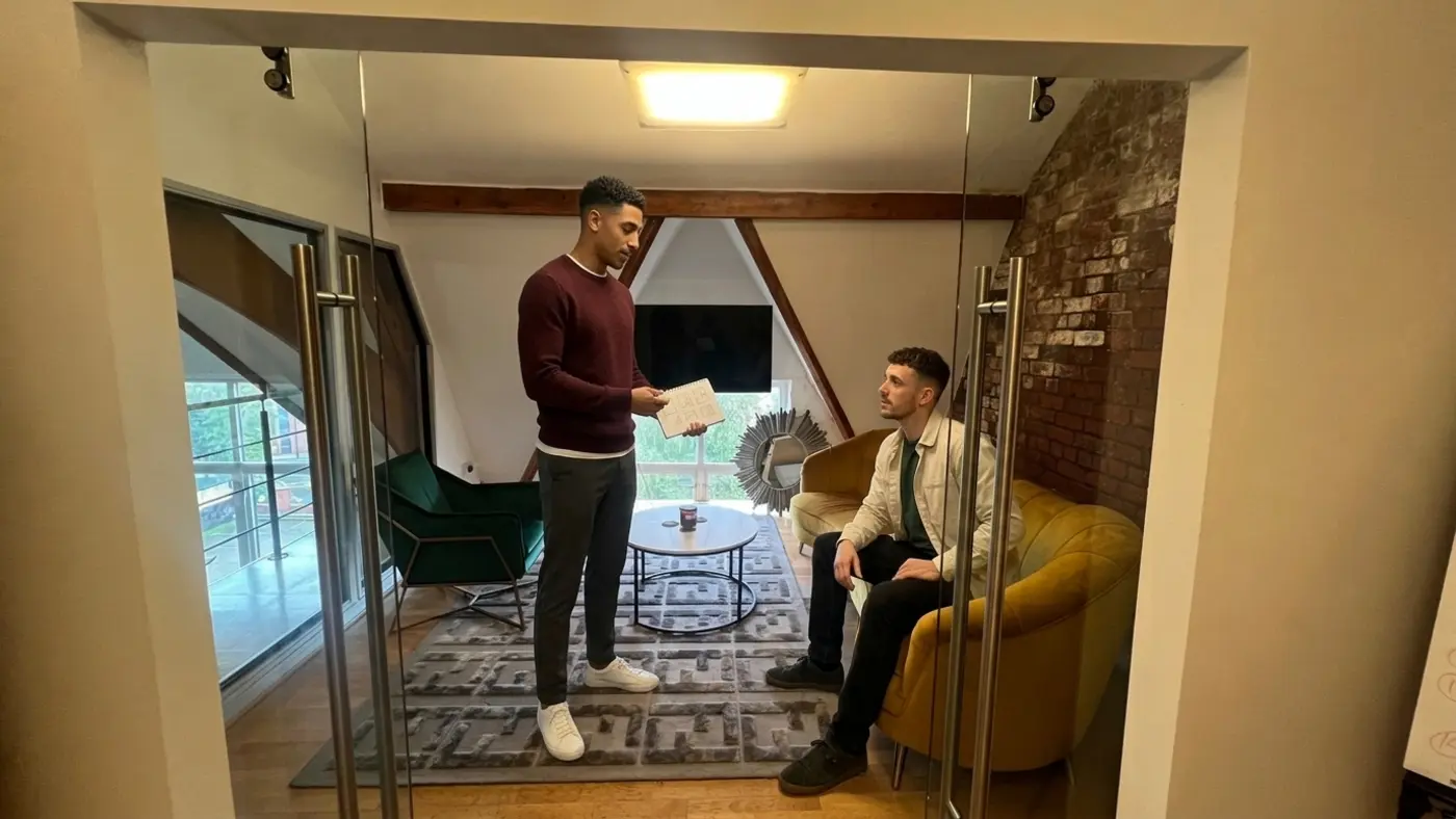 A standing colleague shows a sketchpad to another seated on a sofa in a small modern office