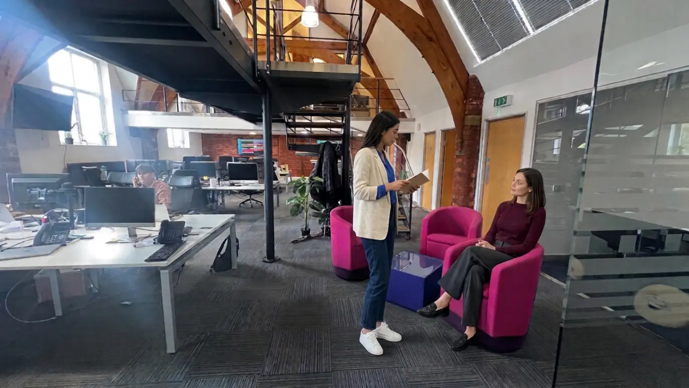 Two colleagues review a notebook by armchairs while a person works at a desk