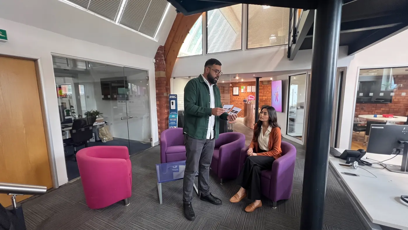Two colleagues compare product idea cards beside purple armchairs in a modern office lobby